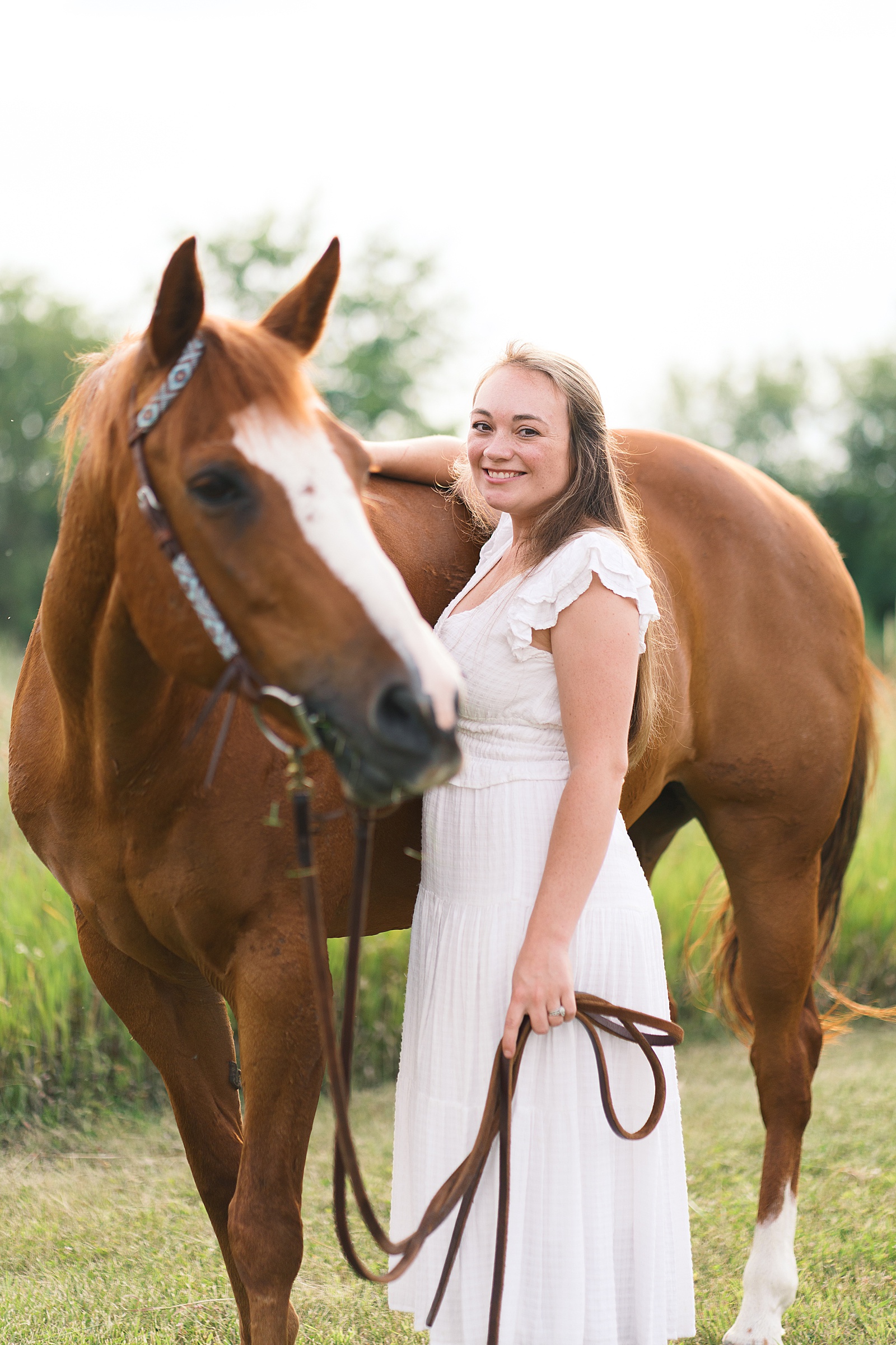 Girl standing at the shoulder of her horse smiling at the camera while her horse looks off in the distance.